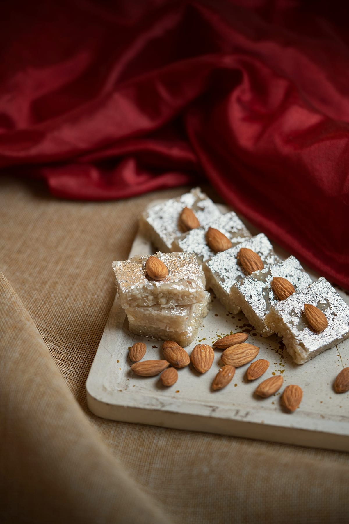 Squares of almond barfi topped with whole almonds on a white board.