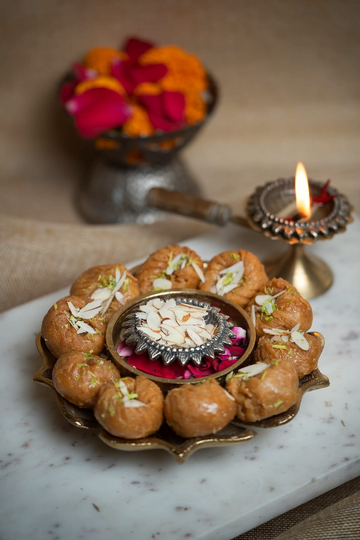 A brass plate ringed with sweets, beside a lit brass lamp and marigolds.
