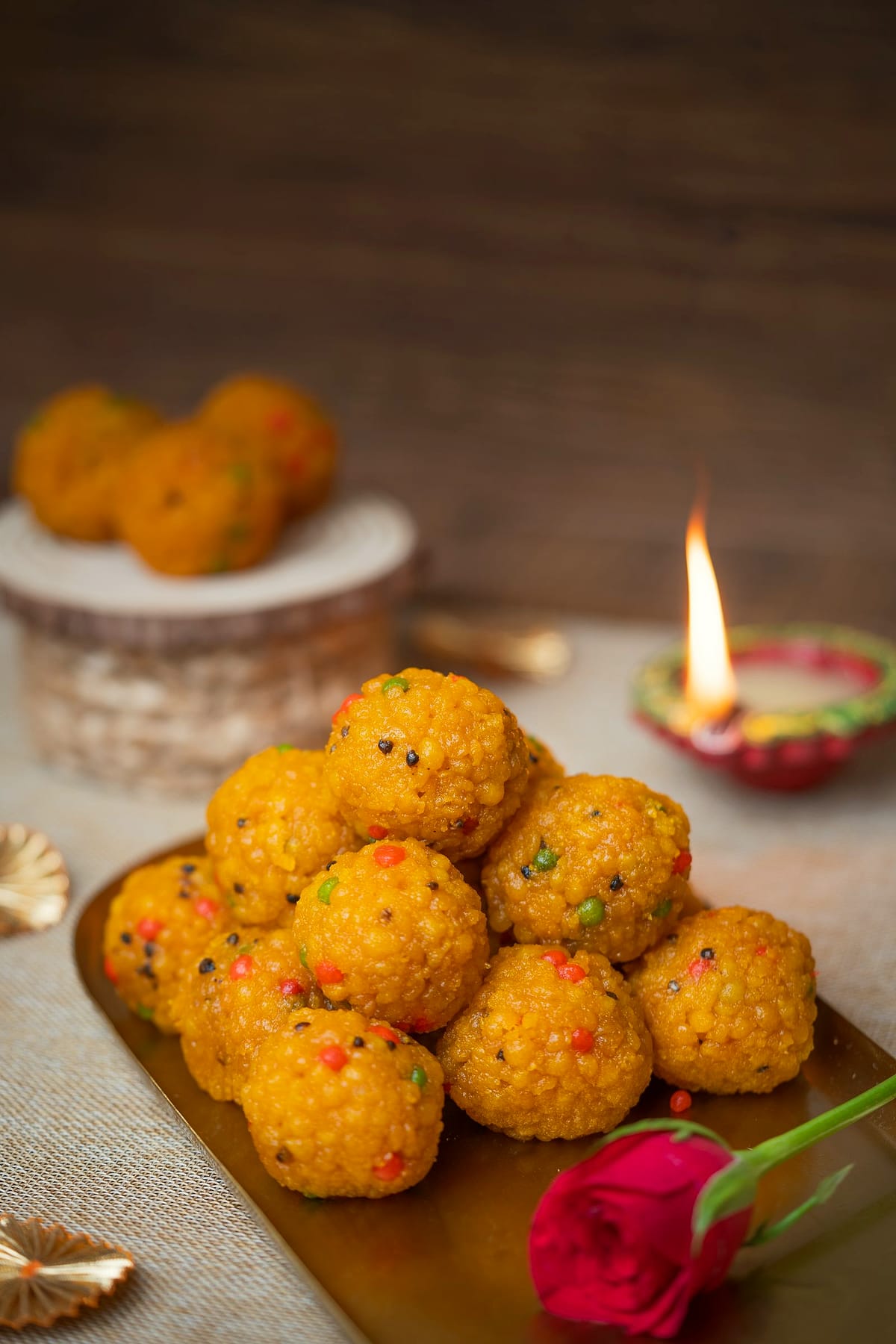 Motichoor laddoos piled on a brass tray beside a lit clay diya and a rose.