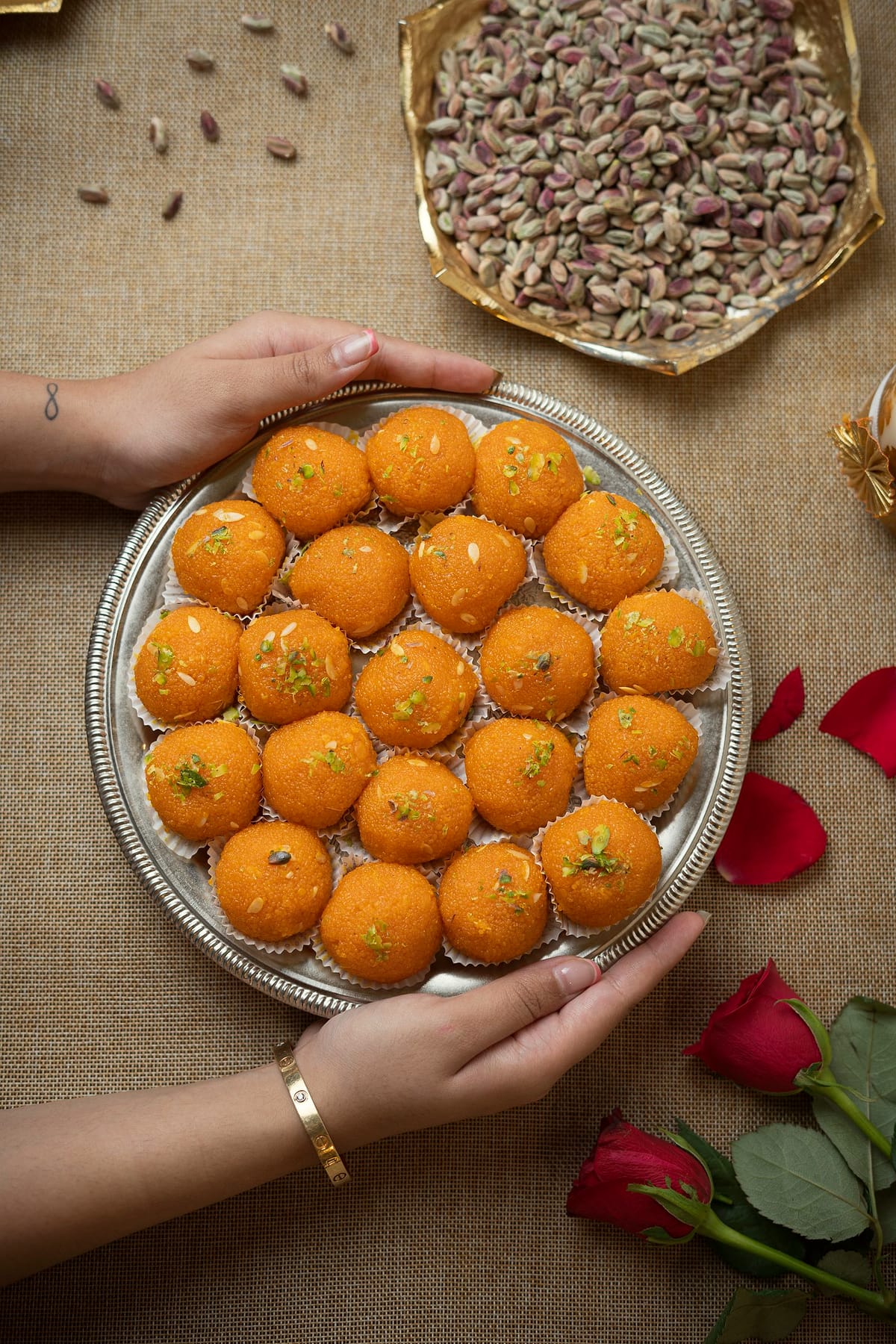 Two hands hold a silver plate of orange boondi laddoos.