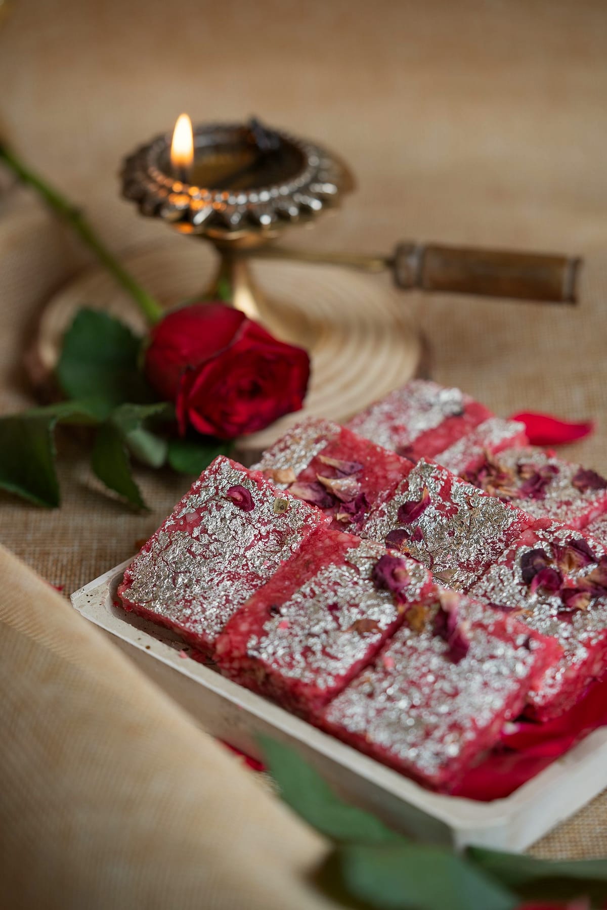 Pink rose barfi with silver leaf on a white tray beside a red rose.